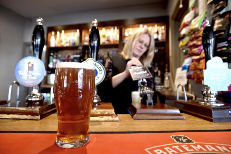 Pint of amber beer on a bar counter in focus, with hand pull taps and drinks display behind, and a person working behind the bar.