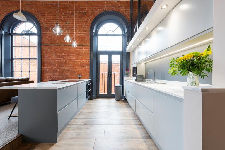 Modern kitchen with exposed brick walls, tall arched windows, and sleek white cabinetry accented by pendant lights and flowers.