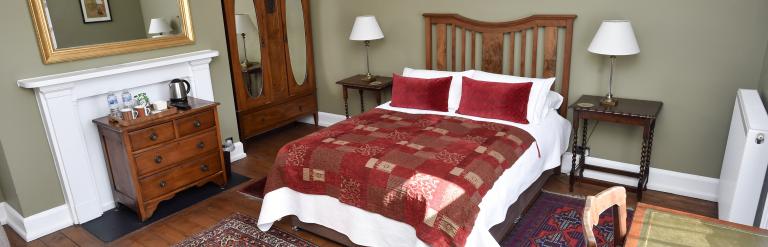 Bedroom with wooden furniture, double bed with red patterned bedding, fireplace mantel, rugs on wooden floor, and a wardrobe and bedside tables against neutral walls.