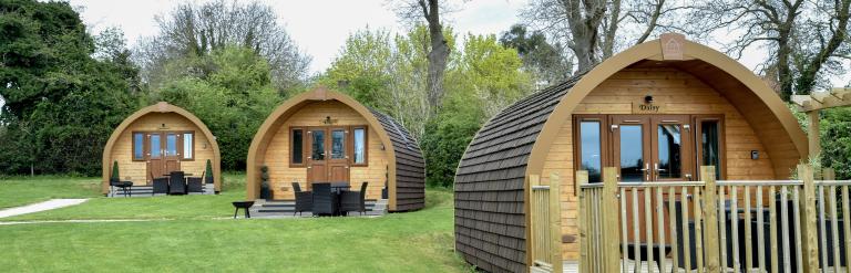 Wooden glamping pods on a grassy site, each with decking and outdoor seating, set among trees along a paved path.