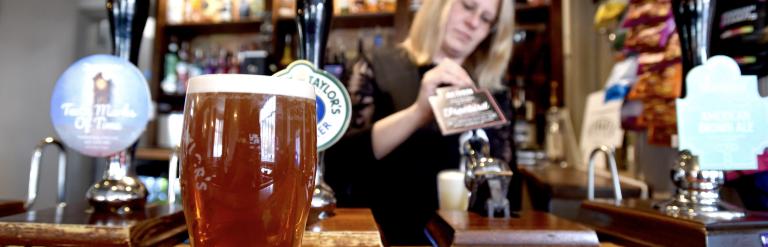 Pint of amber beer on a bar counter in focus, with hand pull taps and drinks display behind, and a person working behind the bar.